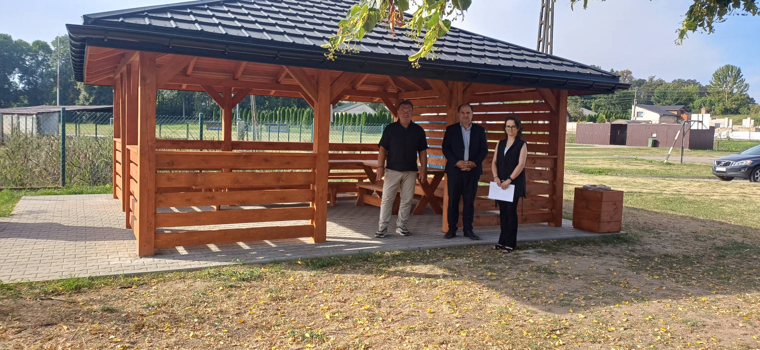 Wójt oraz dwoje pracowników Urzędu Gminy na tle drewnianej altany