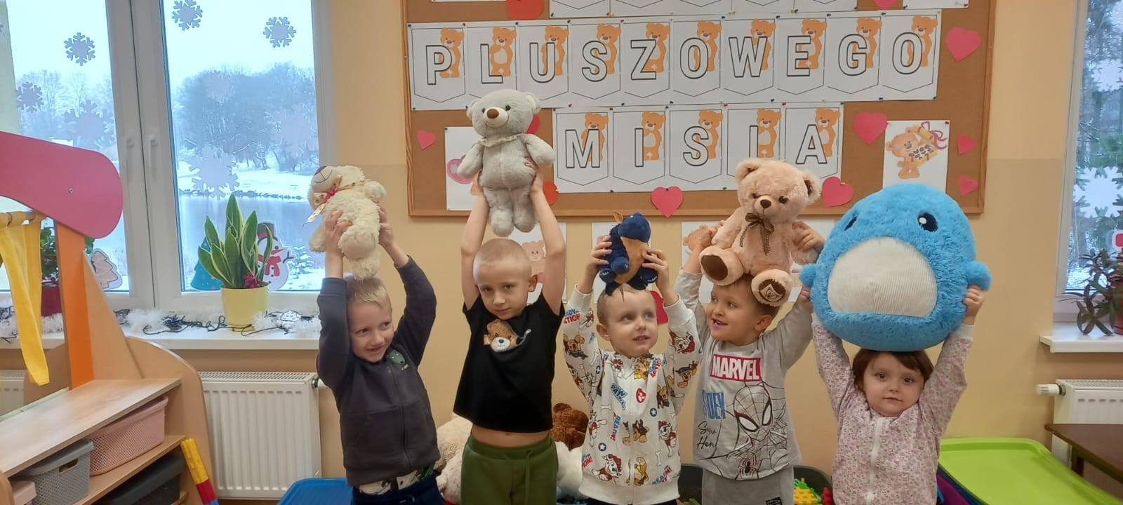 Dzieci trzymają nad sobą pluszowe misie, w tle tablica z częścią napisu pluszowego misia
