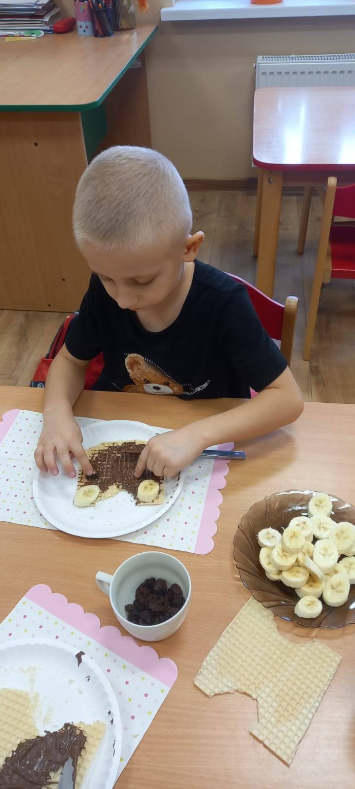 Uczeń siedzący w ławce wykonujący głowę misia z wafelka, czekolady i banana