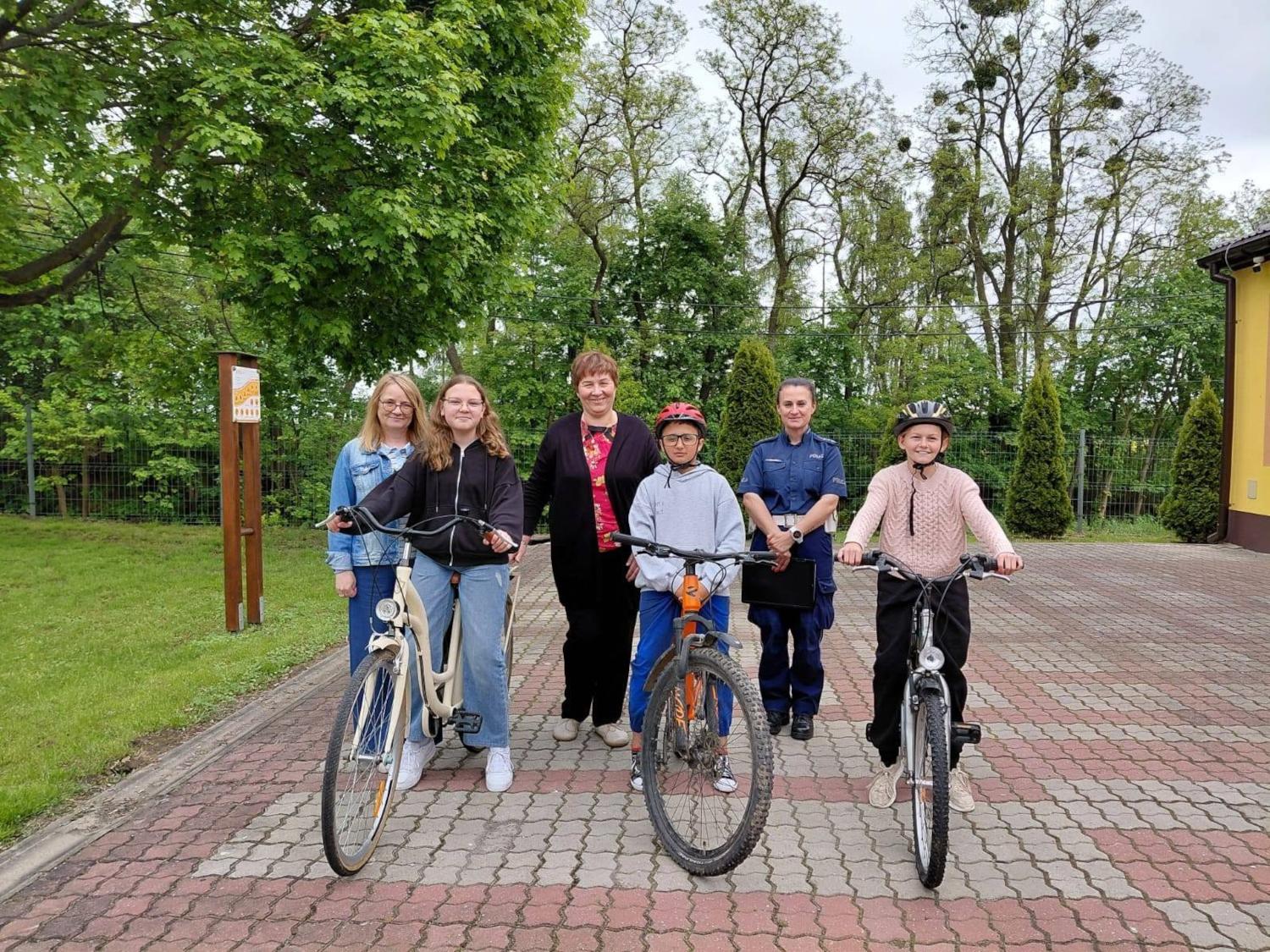 Egzamin na kartę rowerową Dwie dziewczynki i jeden chłopiec siedzą na rowerach, za nimi trzy kobiety, nauczycielka, dyrektor szkoły i policjantka